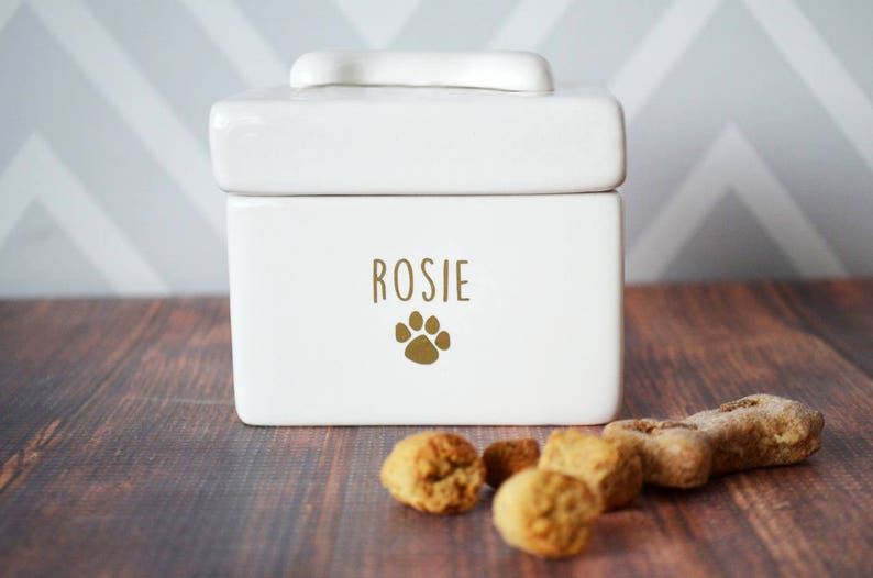 White ceramic dog treat jar with a paw print and the name "Rosie" on the front. The jar has a lid with a bone-shaped handle. There are dog treats on a wooden surface in front of the jar.