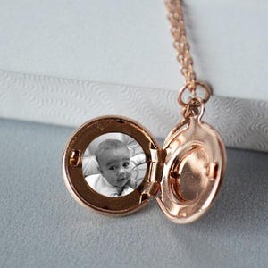 Puede incluir: Un collar con un medallón de oro con una foto de un bebé en su interior. El medallón está abierto, revelando la foto y un espacio en blanco en el otro lado.