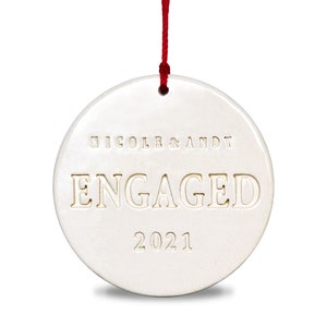 Product image of an off white gloss glazed ceramic round shaped  ornament. The front is stamped with custom text in all capital letters. The text is centered and reads Nicole & Andy, Engaged, 2021. The ornament has a red cord to hang from.