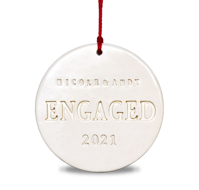 Product image of an off white gloss glazed ceramic round shaped  ornament. The front is stamped with custom text in all capital letters. The text is centered and reads Nicole & Andy, Engaged, 2021. The ornament has a red cord to hang from.