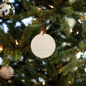 A round shaped, gloss glazed, off-white, ceramic ornament hangs from a red cord on a Christmas tree. The front of the ornament has stamped text, centered and in all capital letters. The tree is decorated with lights and other ornaments.