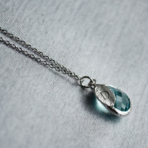 May include: A silver necklace with a teardrop-shaped pendant. The pendant features a faceted, teal-colored gemstone set in a silver leaf-like frame. The chain is delicate and the background is a light gray fabric.