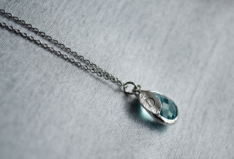 May include: A silver necklace with a teardrop-shaped pendant. The pendant features a faceted, teal-colored gemstone set in a silver leaf-like frame. The chain is delicate and the background is a light gray fabric.