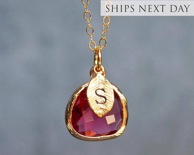 May include: Gold necklace with a red gemstone pendant and a small gold leaf charm with the letter "S" engraved on it.