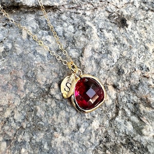 May include: Gold chain necklace with a red gemstone pendant and a small gold leaf charm with the letter 'S' engraved on it.
