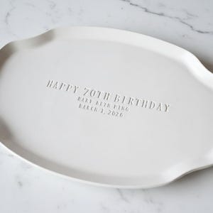 A white ceramic serving platter with a gently scalloped edge. The platter is inscribed with "HAPPY 70TH BIRTHDAY" and the name "Mary Beth Ping" and the date "March 1, 2026".