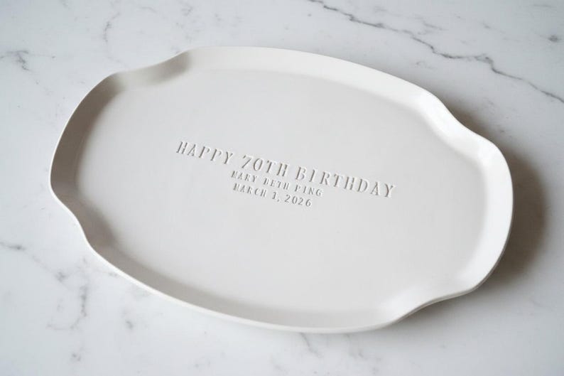 A white ceramic serving platter with a gently scalloped edge. The platter is inscribed with "HAPPY 70TH BIRTHDAY" and the name "Mary Beth Ping" and the date "March 1, 2026".