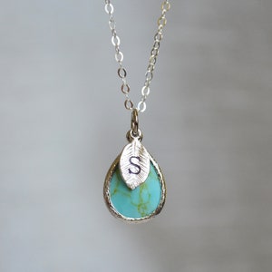 Op de afbeelding: Een zilveren ketting met een turquoise traanvormige hanger en een zilveren blaadje met de letter "S" erop gegraveerd.