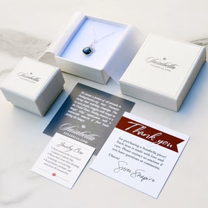 K&ouml;nnte beinhalten: Eine wei&szlig;e Geschenkbox mit einer silbernen Halskette und einem dunkelblauen Edelstein-Anh&auml;nger. Die Box ist mit "Yasabella" und "Susabella.com" beschriftet. Neben der Box liegt eine Dankeskarte mit dem Text "Vielen Dank f&uuml;r den Kauf eines Susabella-St&uuml;cks! Jedes St&uuml;ck wird mit Liebe und Sorgfalt hergestellt. Bitte kontaktieren Sie uns jederzeit, wenn Sie Fragen oder Anmerkungen haben. Mit freundlichen Gr&uuml;&szlig;en! Susan Shapiro".
