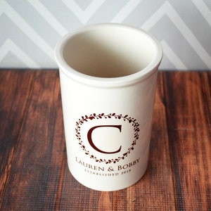 May include: White ceramic utensil holder with a maroon floral wreath design and the text "Lauren & Bobby Established 2018" and the letter "C" in the center.