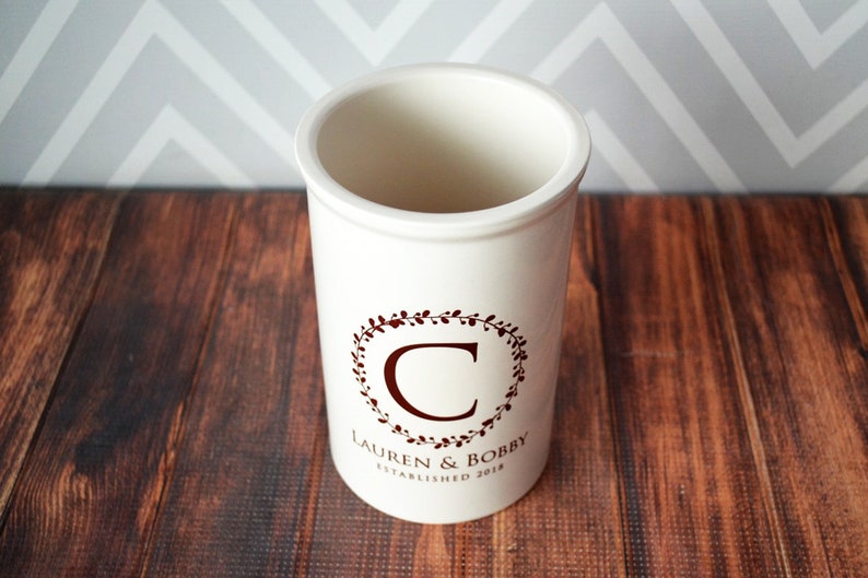 May include: White ceramic utensil holder with a maroon floral wreath design and the text "Lauren & Bobby Established 2018" and the letter "C" in the center.