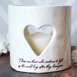 Product image of an off white ceramic votive with a white tea candle inside. The candle is visible through a heart shaped cutout on the front and back. The votive has two lines of centered light brown sepia text below the heart on the front.