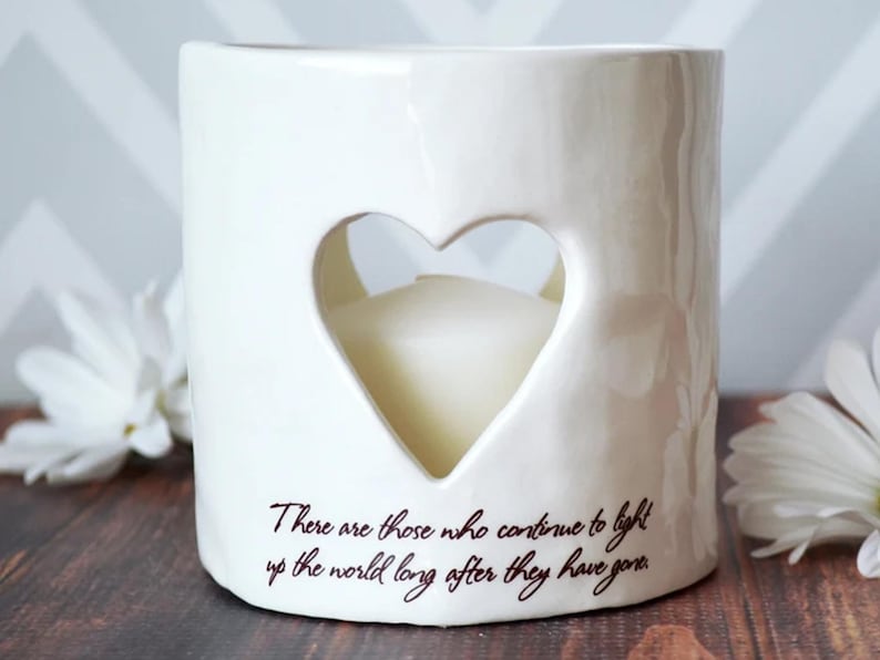 Product image of an off white ceramic votive with a white tea candle inside. The candle is visible through a heart shaped cutout on the front and back. The votive has two lines of centered light brown sepia text below the heart on the front.