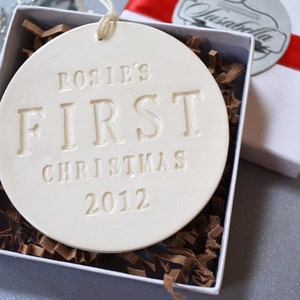 A round shaped, gloss glazed, off-white, ceramic ornament sits in a white gift box on a grey table. The ornament has a white cord to hang from and text stamped into the surface on the front. The text is in all capital letters and centered.