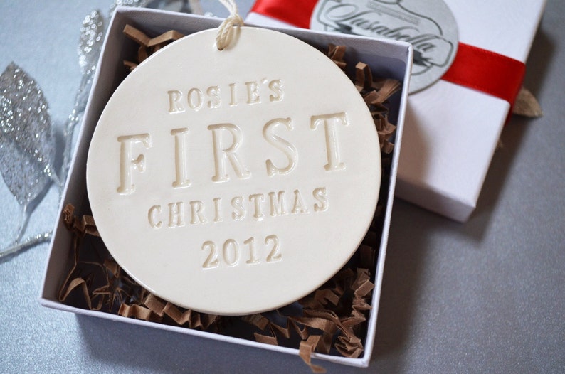 A round shaped, gloss glazed, off-white, ceramic ornament sits in a white gift box on a grey table. The ornament has a white cord to hang from and text stamped into the surface on the front. The text is in all capital letters and centered.