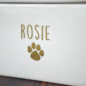 K&ouml;nnte beinhalten: Wei&szlig;e Keramik-Tierfutterdose mit einem goldenen Pfotenabdruck und dem Namen "Rosie" auf der Vorderseite.