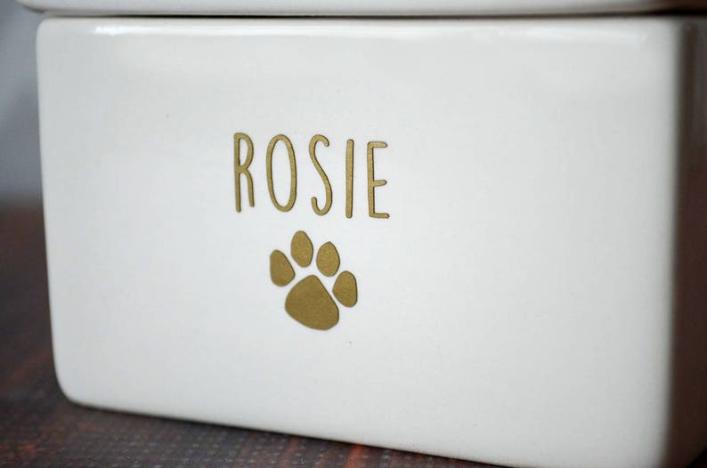 K&ouml;nnte beinhalten: Wei&szlig;e Keramik-Tierfutterdose mit einem goldenen Pfotenabdruck und dem Namen "Rosie" auf der Vorderseite.