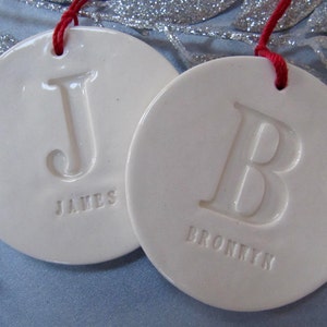 Close up of two off-white, round, ceramic ornaments. Each ornament is stamped with a large initial left unpainted, in white. A small name is stamped below the initial. Each ornament has a red cord to hang it from.