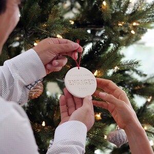 Image of an off white gloss glazed ceramic round shaped  ornament, held in front of a Christmas tree. The front is stamped with custom text in all capital letters. The ornament has a red cord to hang from.