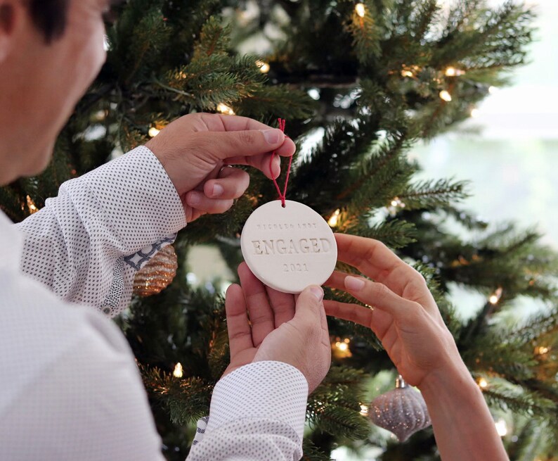 Image of an off white gloss glazed ceramic round shaped  ornament, held in front of a Christmas tree. The front is stamped with custom text in all capital letters. The ornament has a red cord to hang from.