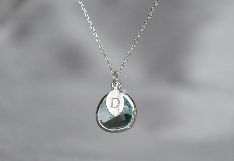 May include: A silver necklace with a teardrop-shaped pendant. The pendant has a light blue gemstone and a silver leaf charm with the letter "D" engraved on it. The chain is delicate and hangs against a blurred background.