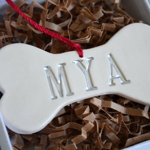 Close up of off-white, ceramic, dog bone-shaped ornament with the pet name "MYA" stamped into the surface. The name is painted in metallic silver. The ornament has a red cord and is shown in a white gift box on silver background.