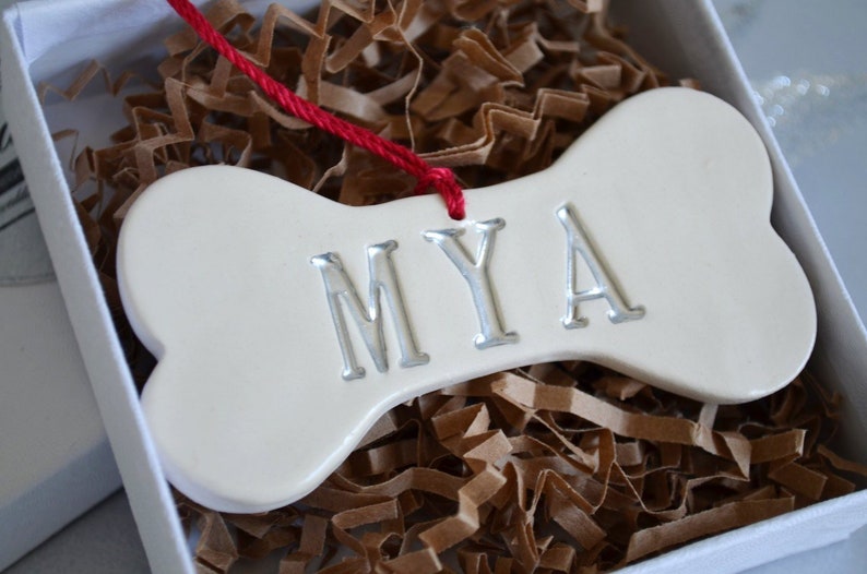 Close up of off-white, ceramic, dog bone-shaped ornament with the pet name "MYA" stamped into the surface. The name is painted in metallic silver. The ornament has a red cord and is shown in a white gift box on silver background.