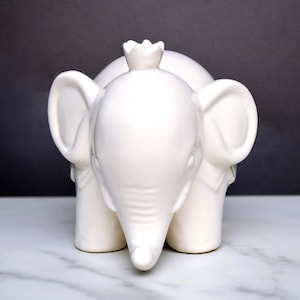 Peut inclure: Figurine d'&eacute;l&eacute;phant en c&eacute;ramique blanche avec une couronne sur la t&ecirc;te. L'&eacute;l&eacute;phant a de grandes oreilles et une trompe, et il repose sur quatre pattes. La figurine est de couleur blanche unie et lisse.