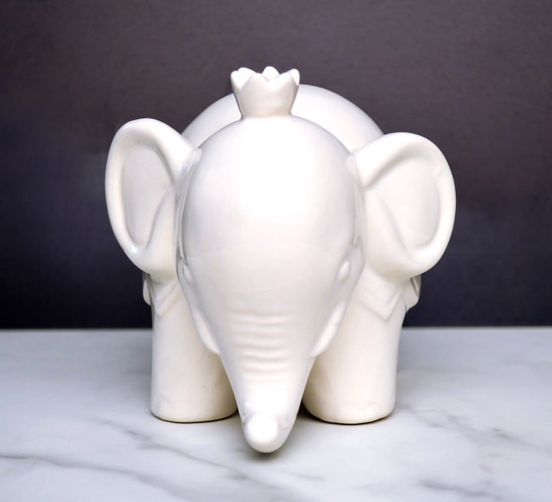 Peut inclure: Figurine d'&eacute;l&eacute;phant en c&eacute;ramique blanche avec une couronne sur la t&ecirc;te. L'&eacute;l&eacute;phant a de grandes oreilles et une trompe, et il repose sur quatre pattes. La figurine est de couleur blanche unie et lisse.