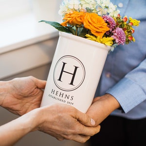 Puede incluir: Un jarr&oacute;n de cer&aacute;mica blanco con la letra "H" dentro de un c&iacute;rculo negro, que contiene un ramo de rosas naranjas, flores moradas y amarillas. El jarr&oacute;n tiene el texto "HEHNS ESTABLISHED 2024".