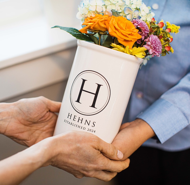 Puede incluir: Un jarr&oacute;n de cer&aacute;mica blanco con la letra "H" dentro de un c&iacute;rculo negro, que contiene un ramo de rosas naranjas, flores moradas y amarillas. El jarr&oacute;n tiene el texto "HEHNS ESTABLISHED 2024".
