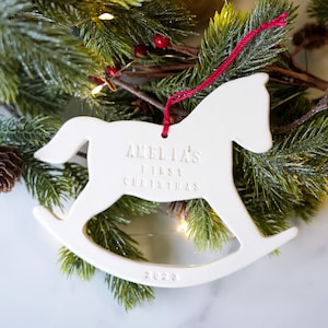 May include: White ceramic rocking horse ornament with the text "Amelia's First Christmas 2023".