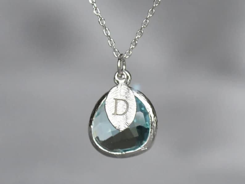 May include: Silver necklace with a teardrop-shaped pendant. The pendant features a light blue gemstone encased in a silver setting. A silver charm with the letter "D" is attached to the pendant. The necklace is on a silver chain.