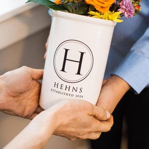 A white ceramic vase with a black circle around the letter 'H' and the words 'Hehns Established 2024' printed on the front. The vase is filled with a bouquet of flowers including orange, yellow, and purple flowers.