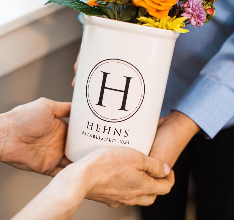 A white ceramic vase with a black circle around the letter 'H' and the words 'Hehns Established 2024' printed on the front. The vase is filled with a bouquet of flowers including orange, yellow, and purple flowers.