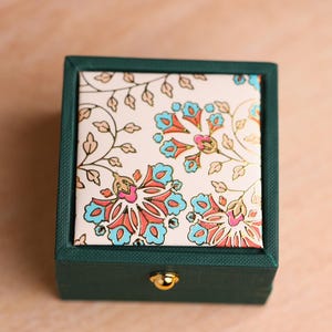 May include: A square jewelry box with a dark green textured base and a lid featuring a cream background with a floral pattern in shades of blue, pink, and gold. A small, round, gold-colored metal knob is centered on the front of the box.
