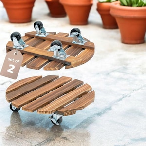 May include: A set of two round wooden plant caddies featuring dark brown stained slats and four black swivel casters each. A light brown tag attached to the top caddy displays the white text set of 2, indicating a pair of mobile plant stands.