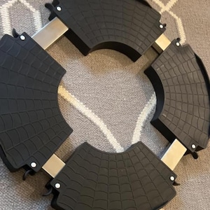 May include: A circular adjustable appliance base featuring four black, textured, fan-shaped corner pieces with a web-like pattern. These pieces are connected by silver-colored metal support bars, forming a sturdy frame designed to hold heavy items.