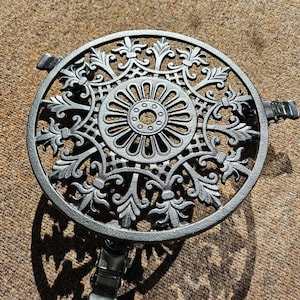 May include: A round black metal plant stand featuring an intricate openwork design with a central circular medallion surrounded by radiating leaf-like patterns. The stand is supported by three short legs, each equipped with a small black caster wheel.