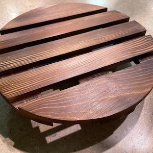 May include: A circular wooden stand constructed from parallel dark brown stained planks with visible wood grain. The design features evenly spaced gaps between the slats, creating a slatted surface with a rounded edge profile and a sturdy base.