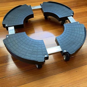 May include: A circular adjustable plant caddy featuring four black plastic corner segments with a textured spiderweb pattern, connected by silver metal support bars, and equipped with small swivel wheels attached to the underside of each corner.