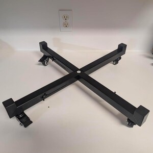 May include: A black, cross-shaped adjustable stand featuring four sturdy arms with raised end caps. The base is equipped with four small, swiveling caster wheels, each including a locking mechanism to secure the stand in a fixed position.