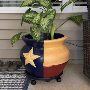 May include: A round ceramic planter featuring a painted design inspired by the Texas flag, with a blue section containing a large yellow star, a white middle section, and a red bottom section, resting on a black circular plant caddy with wheels.