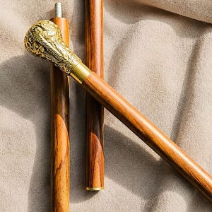 May include: A walking stick featuring a polished gold-tone metal handle with intricate embossed floral patterns. The shaft is constructed from brown wood with visible grain, and the segments are connected by threaded metal joints with a rubber tip.