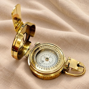 May include: A polished brass pocket compass with a hinged lid and a circular dial featuring cardinal directions, degree markings, and a central needle. The device includes a small metal loop on the side and a clear glass cover over the internal scale.