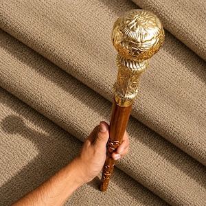 May include: A walking stick featuring a polished gold-tone spherical handle with intricate embossed floral and geometric patterns. The shaft is crafted from smooth brown wood, showcasing carved decorative details along its length.