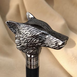 May include: A walking stick handle shaped like a wolf head, crafted from polished silver-toned metal with detailed fur texture and pointed ears. The handle is mounted on a smooth, dark-colored shaft with a metallic collar featuring horizontal lines.