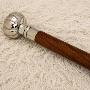 May include: A walking stick featuring a polished silver-tone metal handle with a rounded, ribbed pumpkin-like shape. The handle connects to a smooth, dark brown wooden shaft via a cylindrical silver-tone metal collar, creating a sleek design.