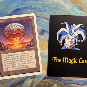 May include: A Magic: The Gathering Chaos Orb trading card featuring a brown border and an illustration of a floating orb over lava, alongside a black rectangular card displaying a jester illustration and the yellow text The Magic Lair.