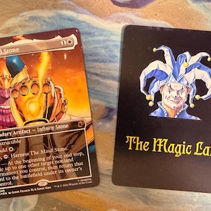 May include: Two rectangular trading cards. The left card features Thanos holding a glowing golden gauntlet with the text The Mind Stone, Legendary Artifact, and Infinity Stone. The right card is black with a jester illustration and The Magic Lair text.